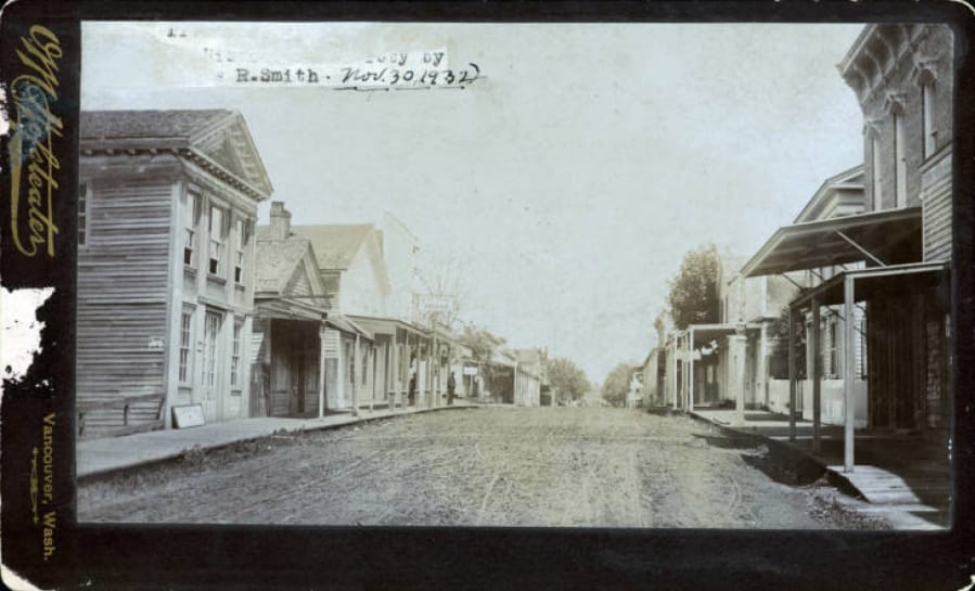 This view looks north on Main Street Vancouver in 1882. On the left, a sign for the Pacific House juts out. In the distance, two men loiter on the boardwalk. A little farther west of the Pacific House, the former city commons was slowly transforming into Esther Short Park.