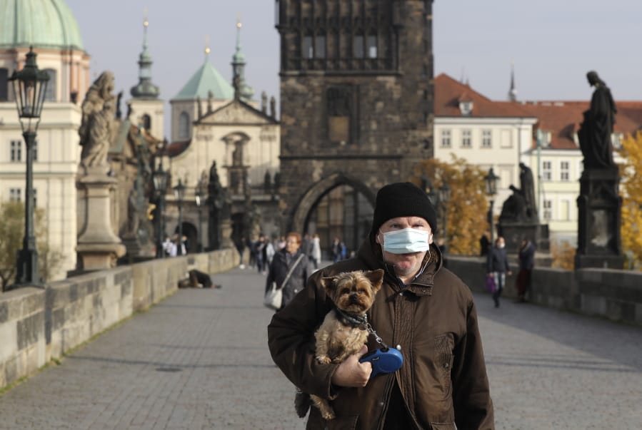 A man wearing a face mask carries his dog across the medieval Charles Bridge in Prague, Czech Republic, Wednesday, Oct. 21, 2020. In another desperate attempt to slow the rise of coronavirus infections in the Czech Republic, Health Minister Roman Prymula has announced a ban on free movement of people in the country and a closure of many stores, shopping malls and hotels. At the same time, state offices will limit their opening hours. Prime Minister Andrej Babis says those measures should prevent the collapse of the health system in early November.