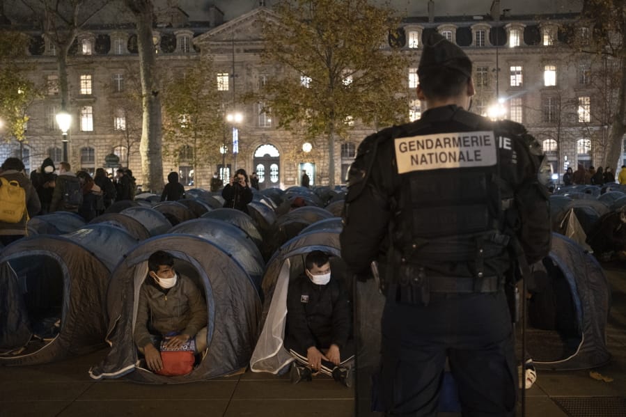 A police officer stands in front of migrants in a makeshift camp set up Monday night Nov.23, 2020 on Place de la Republique in Paris. Paris police are under government orders to explain themselves after officers were filmed tossing migrants out of tents while evacuating a makeshift camp in the French capital.
