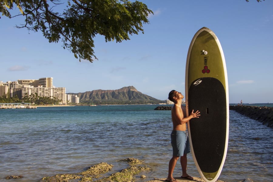 In this photo provided by Yoko Liriano, Bryant de Venecia poses for a photo with his paddleboard in Honolulu, Wednesday, Nov. 11, 2020. He started stand-up paddle-boarding when there were fewer tourists coming to Hawaii during the pandemic. He&#039;s among the Hawaii residents feeling ambivalence toward tourists returning now that the state is allowing incoming travelers to bypass a 14-day quarantine with a negative COVID-19 test.