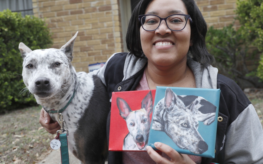 In this photo made Monday, March 2, 2020, Danielle Moore poses for photos with Kana and also paintings of the pet Australian cattle dog in Dallas. In the dog-eat-dog world of online shopping, Chewy has an unusual plan to fend off Amazon: turning pets into works of art. The online pet shop surprises customers with oil paintings of their furry friends, a move the company says wins them customers for life.
