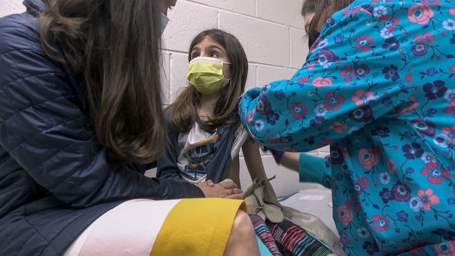 In this Wednesday, March 24, 2021 image from video provided by Duke Health, Alejandra Gerardo, 9, looks up to her mom, Dr. Susanna Naggie, as she gets the first of two Pfizer COVID-19 vaccinations during a clinical trial for children at Duke Health in Durham, N.C. In the U.S. and abroad, researchers are beginning to test younger and younger kids, to make sure the shots are safe and work for each age.