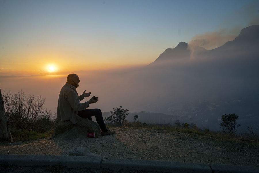 A Capetonian watches the sun rise from the top of Signal Hill as smoke engulfs the city of Cape Town, South Africa, Tuesday April 20, 2021. A massive fire spreading on the slopes of the city's famed Table Mountain, at right, is kept under control as firemen and helicopters take advantage of the low winds to contain the blaze.