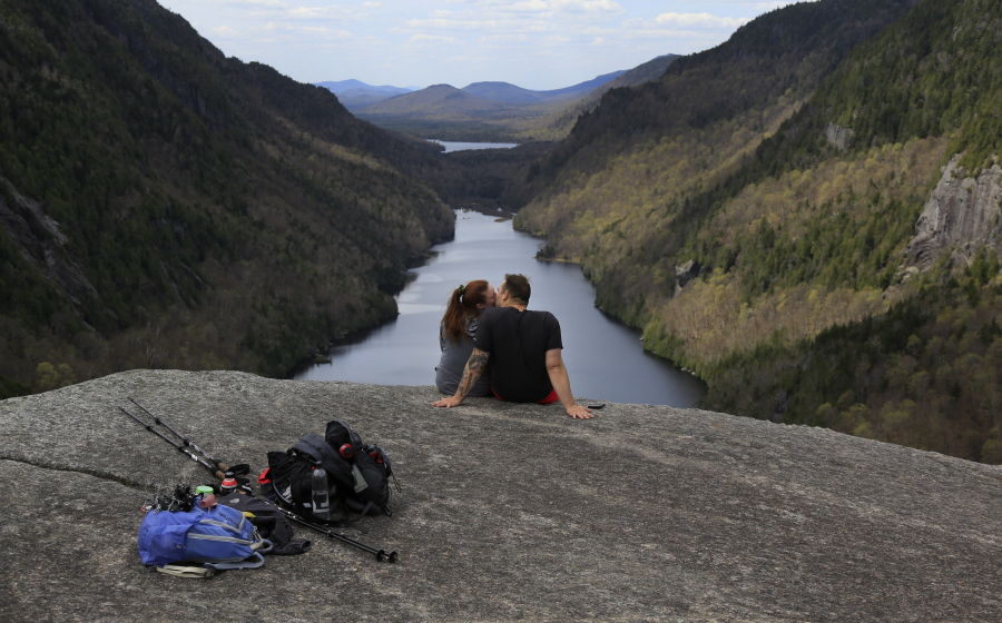 Sidney Gleason, left, and Joe Gorsuch, both of Syracuse, kiss while taking in the view of Lower Ausable Lake at Indian Head summit inside the Adirondack Mountain Reserve, Saturday, May 15, 2021, near St. Huberts, N.Y. A free reservation system went online recently to control the growing number of visitors packing the parking lot and tramping on the trails through the private land of the Adirondack Mountain Reserve. The increasingly common requirements, in effect from Maui to Maine, offer a trade-off to visitors, sacrificing spontaneity and ease of access for benefits like guaranteed parking spots and more elbow room in the woods.