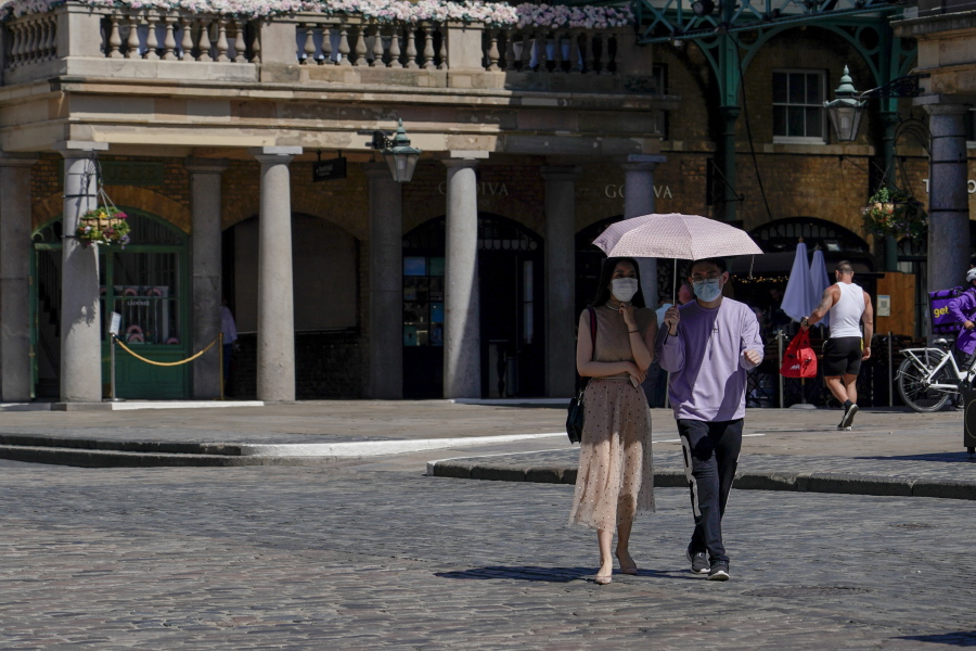 A couple wear face masks as they shelter from the sun under an umbrella, while they walk in Covent Garden, in London, Monday, June 14, 2021. British Prime Minister Boris Johnson is expected to confirm Monday that the next planned relaxation of coronavirus restrictions in England will be delayed as a result of the spread of the delta variant first identified in India.