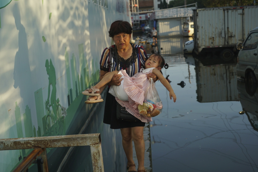 A woman carries a child in her arms as she walks on a curb above floodwaters in Xinxiang in central China's Henan Province, Monday, July 26, 2021. Record rain in Xinxiang last week left the produce and seafood market soaked in water. Dozens of people died in the floods that immersed large swaths of central China's Henan province in water.