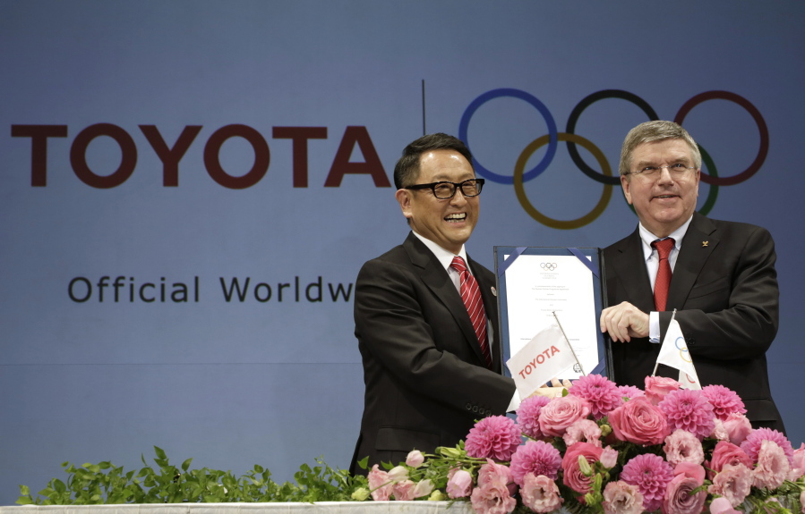FILE - In this March 13, 2015 file photo, Toyota President and CEO Akio Toyoda, left, and IOC President Thomas Bach pose with a signed document during a press conference in Tokyo as Toyota signed on as a worldwide Olympic sponsor in a landmark deal, becoming the first car company to join the IOC's top-tier marketing program. Toyota won't be airing any Olympic-themed advertisements on Japanese TV during the Tokyo Games despite being one of the IOC's top corporate sponsors.