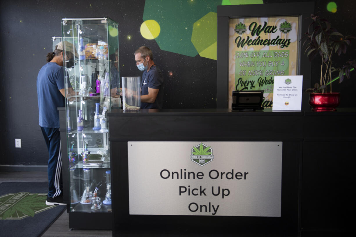 Budtender/cashier Robert Emmons, in blue mask, assists a customer as they pick up an online order inside Main Street Marijuana in Uptown Village on Tuesday morning. The store has put up new signs advising customers that all online orders must now be picked up at indoor counters following a change to state rules three weeks ago.