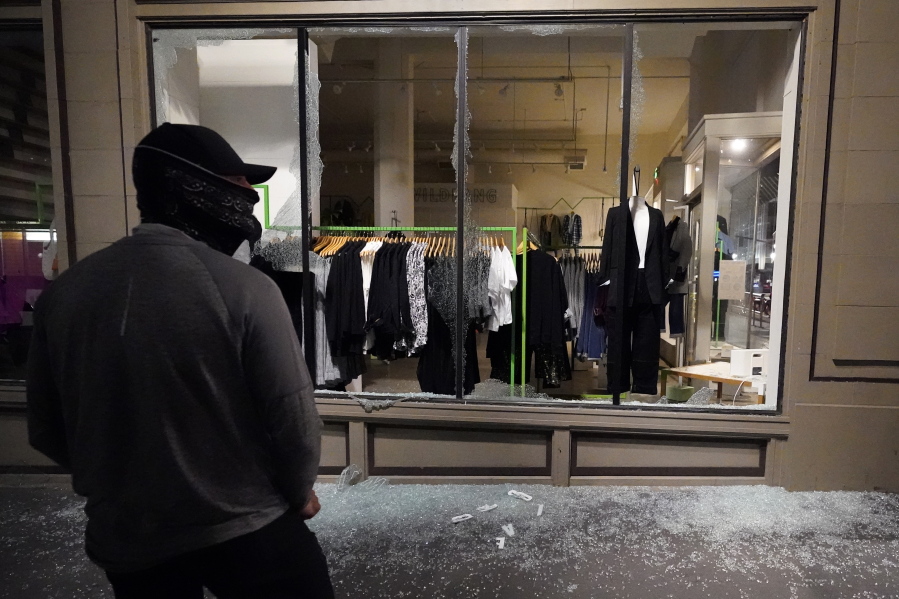 FILE - In this Nov. 4, 2020, file photo, a man stands in front of a broken display window at a retail store during protests in Portland, Ore. Police in Portland say they believe a new state law prohibits officers from directly intervening when people smash storefronts and cause mayhem.