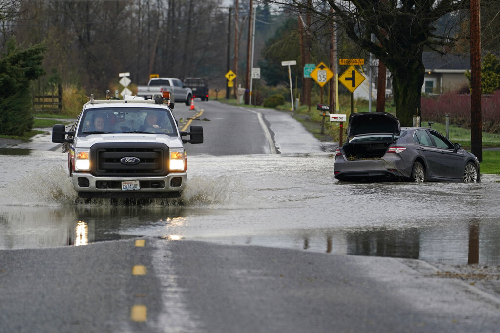 A truck drives through water over a road near Everson, Wash., Monday, Nov. 29, 2021 past a car that was stranded by flooding in the area earlier in the month. Localized flooding was expected Monday in Washington state from another in a series of rainstorms, but conditions do not appear to be as severe as when extreme weather hit the region earlier in November.