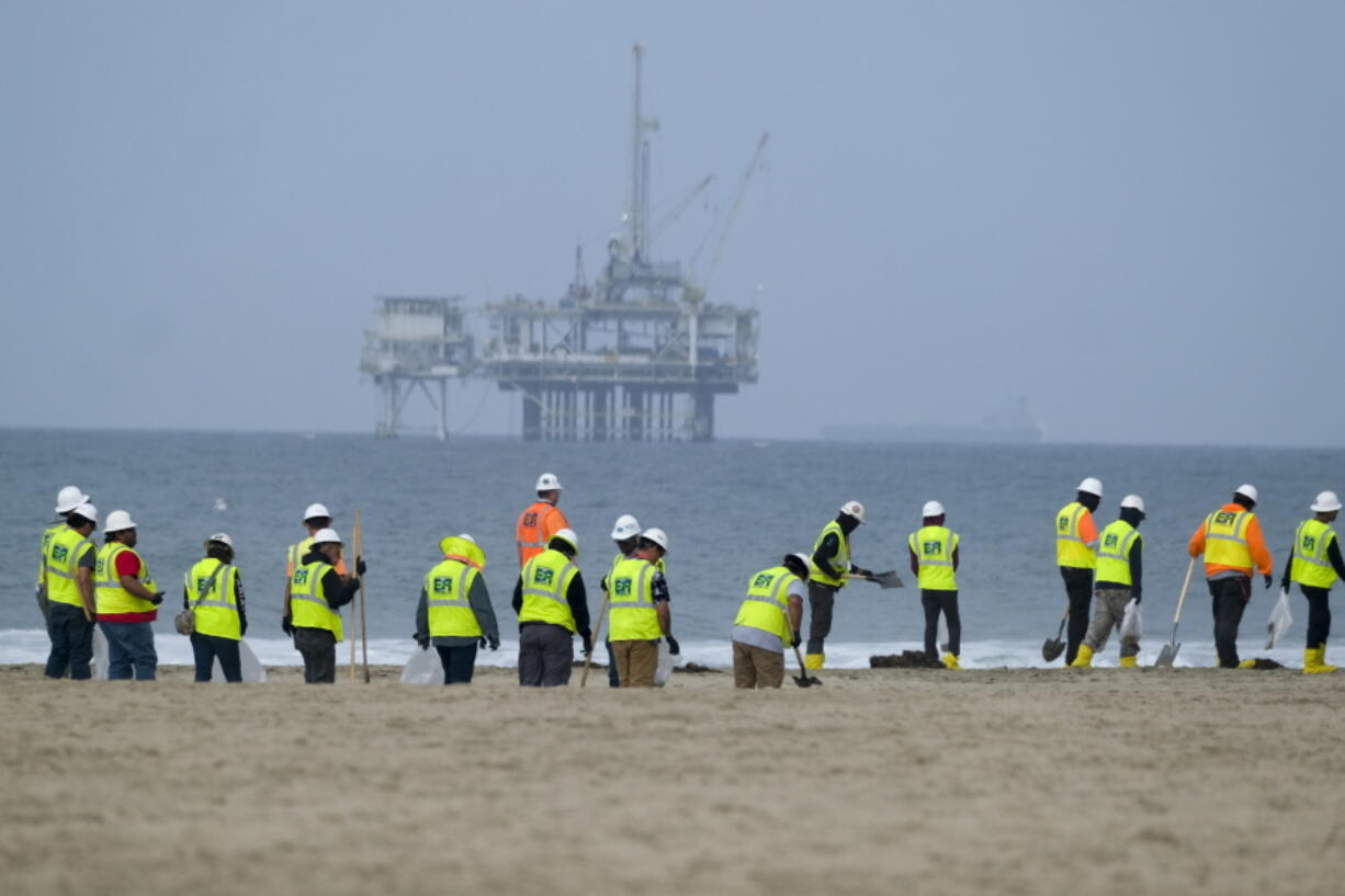 FILE - Workers in protective suits continue to clean the contaminated beach with a platform in the background in Huntington Beach, Calif., on Oct. 11, 2021. A month after an offshore oil spill, environmental advocates say they plan to sue the federal government over the failure to review and update plans for platforms off the Southern California coast. The Center for Biological Diversity sent a notice to the Secretary of the Interior Tuesday, Nov. 2, 2021, of its intent to sue. (AP Photo/Ringo H.W.