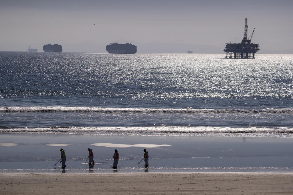 Container ships and an oil derrick line the horizon as environmental oil spill cleanup crews search the beach, cleaning up oil chucks from a major oil spill in Huntington Beach Tuesday, Oct. 5, 2021. (Allen J.