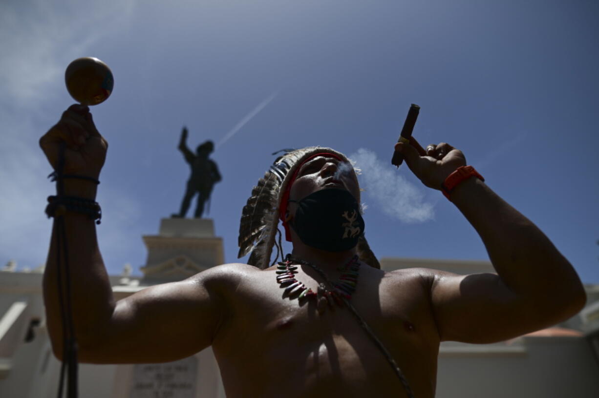 FILE - Gypsy Cordova blows smoke and shakes a maraca in front of a Juan Ponce de Leon monument while leading a group of activists in a march demanding statues and street names commemorating symbols of colonial oppression be removed, in San Juan, Puerto Rico, Saturday, July 11, 2020. Unknown people have toppled a statue of Spanish explorer Juan Ponce de Le?n ahead of a visit of King Felipe VI to the U.S. territory of Puerto Rico. Col. Jos? Juan Garc?a, police commissioner for San Juan, told The Associated Press  on Monday, Jan. 24, 2022 that officers patrolling the cobblestone streets of the capital's historic district hear a loud bang at 4:30 a.m. and found the statue broken in pieces.