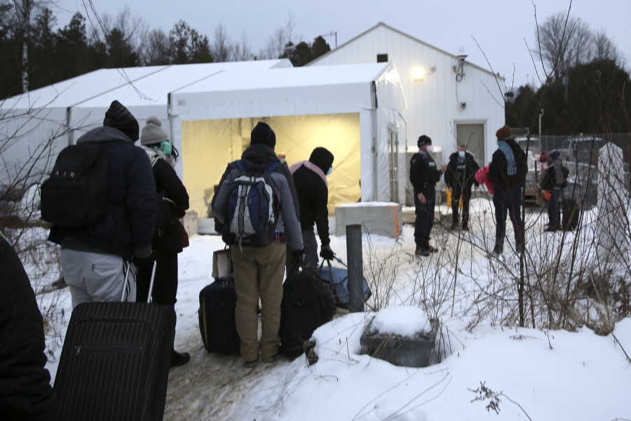 Migrants line up on the border of the United States, foreground, and Canada, background, at a reception center for irregular borders crossers, in Saint-Bernard-de-Lacolle, Quebec, Canada, Wednesday Jan. 12, 2022, in a photo taken from Champlain, N.Y. They are crossing the U.S.-Canadian border into Saint-Bernard-de-Lacolle, Quebec, where they are arrested by the Royal Canadian Mounted Police and then allowed to make asylum claims. The process was halted for most cases after the 2020 outbreak of COVID-19, but the Canadian government changed its policy in November, allowing the process to continue.