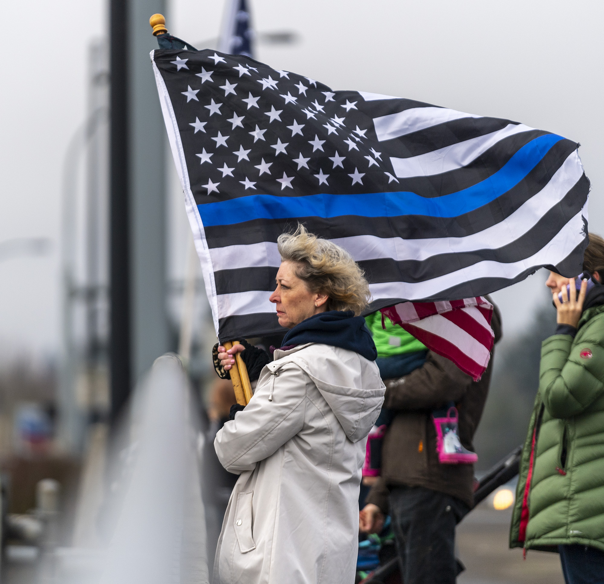 Tricia Davis of Battle Ground holds a thin blue line flag at the Pioneer Street I-5 overpass in Ridgefield on Tuesday, Feb. 8, 2022, before a funeral processional for Vancouver Police Department Officer Donald Sahota, who was fatally shot Jan. 29 during a manhunt for an armed robbery suspect.