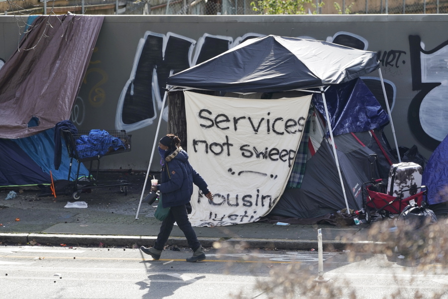 A person walks past a tent used by people experiencing homelessness with a sign on it that reads "services not sweeps," Tuesday, March 1, 2022, in downtown Seattle, across the street from City Hall. For years, liberal cities in the U.S have tolerated people living in tents in parks and public spaces, but increasingly leaders in places like Portland, Oregon, New York and Seattle are removing encampments and pushing other strict measures that would've been unheard of a few years ago. (AP Photo/Ted S.