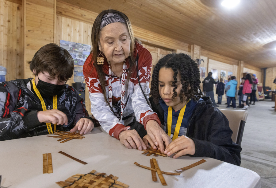 Grace Hyasman, center, a canoe journey specialist with the Nisqually tribe, helps Christian Jensen, 10, learn how to weave a mat during a workshop during his visit to the Nisqually Cultural Center in DuPont Tuesday, April 26, 2022. He was there with other fourth graders from the North Thurston School District. At left is Jordan James, 10. They are making mats in the Longhouse at the center. (Ellen M.