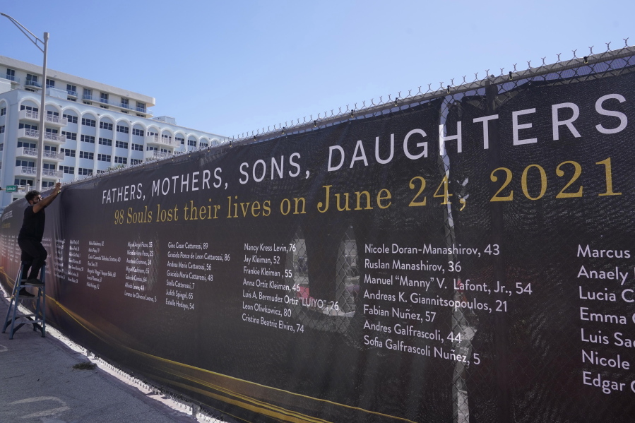 Christopher Rosa Cruz installs a large banner at the site of the Champlain Towers South condominium building, Thursday, May 12, 2022, in Surfside, Fla. The banner lists the names of the 98 people killed when the building suddenly collapse nearly a year ago.