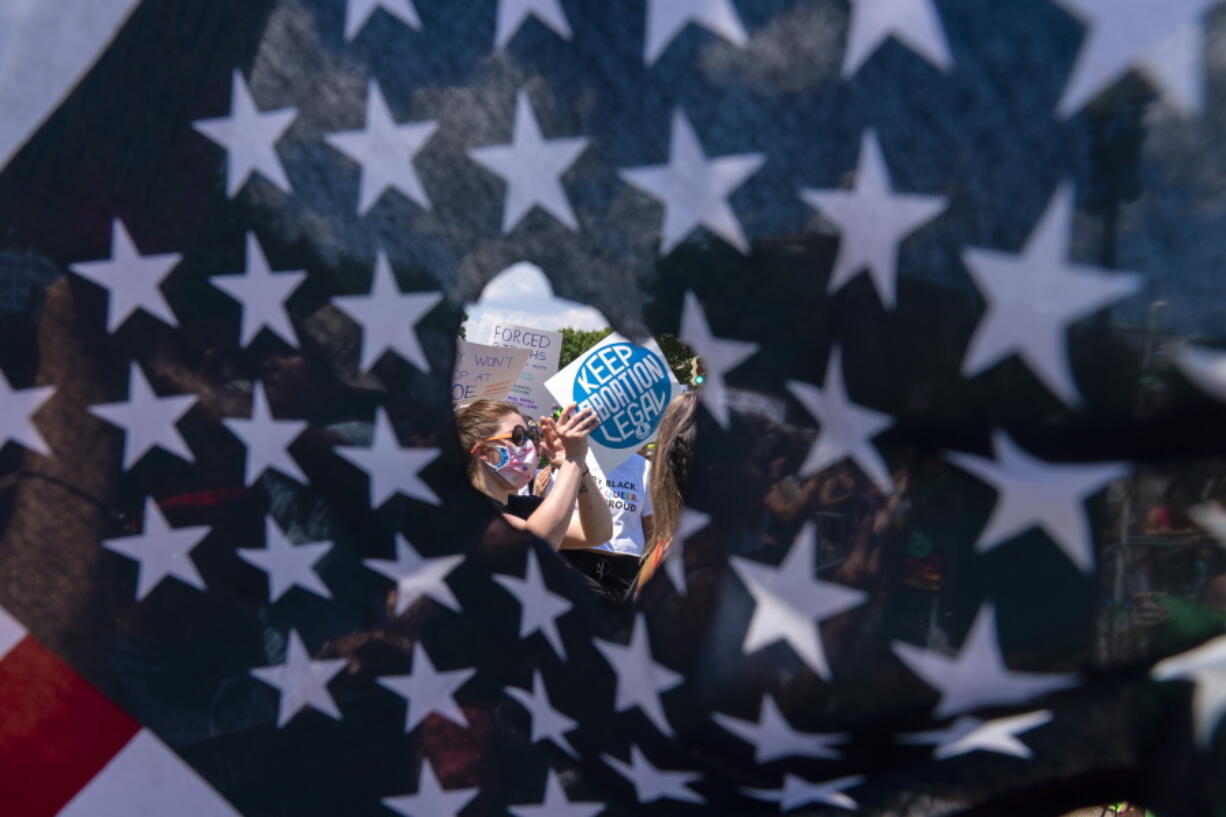 Abortion rights activists are seen through a hole in an American flag as they protest outside the Supreme Court in Washington, Saturday, June 25, 2022. The Supreme Court's decision to overturn national protections for abortion has set off a contest between Democratic and Republican lawmakers over whose policies would do more to help vulnerable mothers and children. It's a key issue going into the midterm elections.