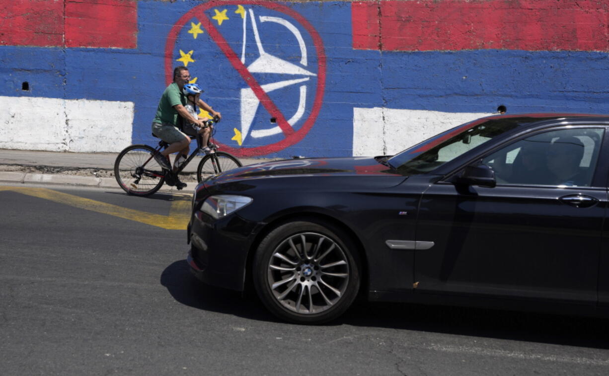 A man with a child rides a bicycle by a wall that shows the colours of the Serbian flag and graffiti against EU and NATO in Belgrade, Serbia, Tuesday, June 21, 2022. European Union leaders will seek to offer support this week to six Western Balkan nations that have long been knocking at the bloc's doors, and now see the war in Ukraine raging not far from their borders amid fears that Russia could turn its sights on their region. Serbia, the largest of the six countries, is pivotal in Russia's regional influence.