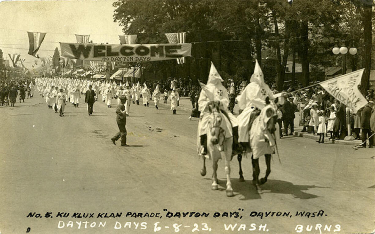 Black and white postcard photograph of a large group of robed and hooded Ku Klux Klan members participating in the Dayton Days parade, Dayton, Columbia County, WA, June 8, 1923. It is led by two Klan members on horses with drapes, and carrying a banner reading "Dayton/Ku Klux Klan." They are followed by a large number of men in Klan robes and hoods, perhaps more than 100, probably members of the local Klaven. A man wearing a suit leads this group. A large banner over the street reads, "Welcome/ to Dayton Days." American flags and another banner are also over the street. Many spectators are on both sides of the street, including small girls in image right.