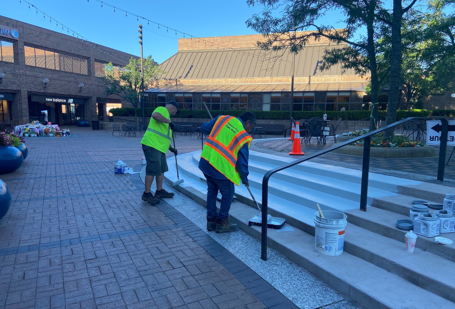 This image provided by Nancy Schneider shows workers painting steps in a business district that reopened Sunday, July 10, 2022 in Highland Park, Ill. It had been closed as a crime scene since the July 4 mass shooting that killed seven people. In the background is a makeshift memorial to victims.