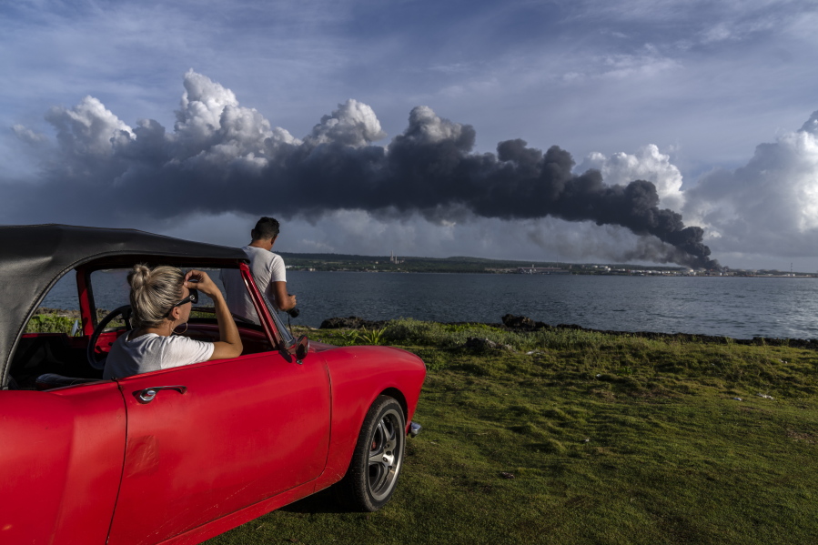 People watch a huge plume of smoke rise from the Matanzas supertanker base, as firefighters work to douse a fire that started during a thunderstorm the night before, in Matanzas, Cuba, Sunday, Aug. 7, 2022. Cuban authorities say lightning struck a crude oil storage tank at the base, sparking a fire that sparked four explosions that injured more than 121 people, one person dead and 17 missing.