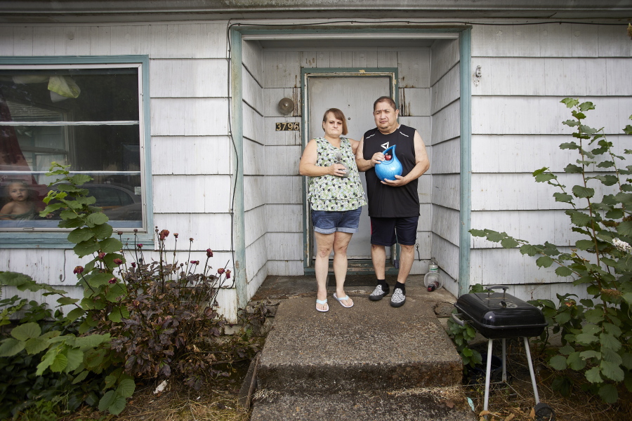 Misty Castillo, left, and her husband Arcadio stand in front of their home, holding the urn containing the ashes of their son Arcadio Castillo III, who was shot in 2021 by Salem police, as their granddaughter Nala, age 2, looks out the window in Salem, Ore., Thursday, Aug. 18, 2022. Misty called 911 and asked for the police, saying her son was mentally ill, was assaulting her and her husband and had a knife. Less than five minutes later, a police officer burst into the house and shot him.