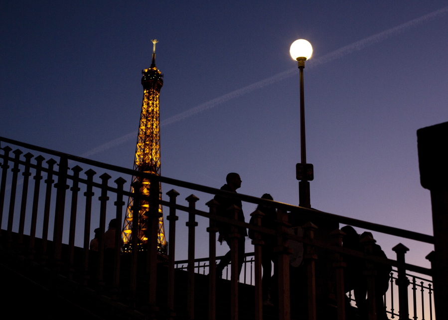 FILE - People walk on a bridge next to the Eiffel Tower in Paris, Wednesday Feb. 9, 2022. Lights on the Eiffel Tower will soon be turned off an hour earlier at night as part of an energy savings plan in the French capital, its mayor announced. Paris mayor said the iconic tower that is illuminated until 1:00am is only one of the city's monuments and municipal buildings that will be plunged into darkness earlier in the evening as the French capital faces risks of power shortages, rationing and blackouts when energy demand surges this winter.