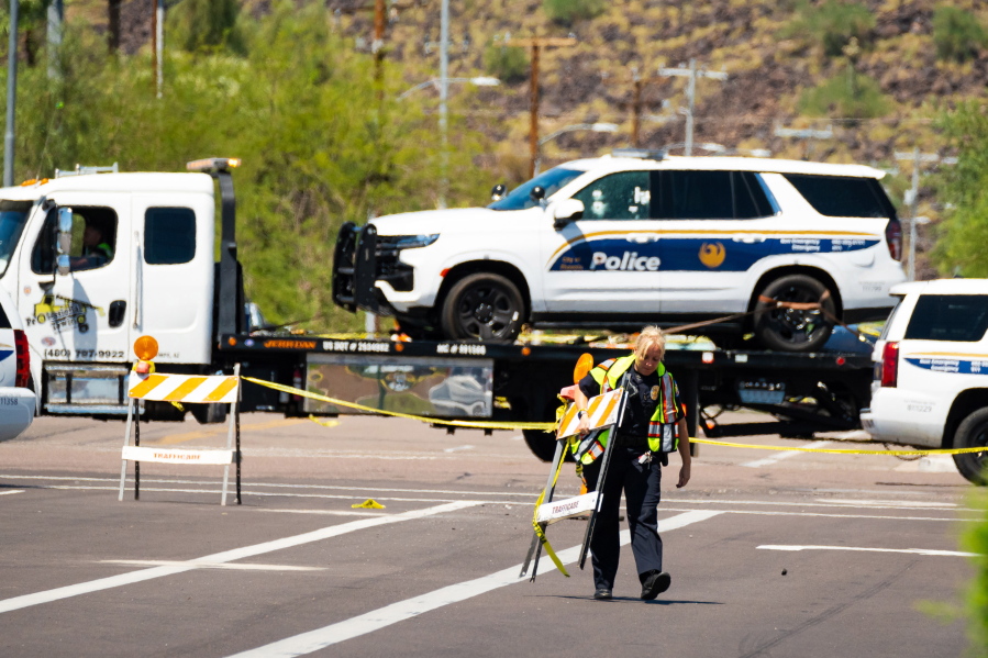 FILE - A police officer carries traffic barricades across 27th Avenue near Dear Valley Road, Sunday, Aug. 29, 2022, in Phoenix, as a damaged police vehicle is transported out of the area after a shooting occurred the night before injuring two Phoenix police officers. Americans struggled this week to process not one, but multiple high-profile shootings that unfolded in major cities and smaller towns across the U.S. But behind the stand-out headlines about shooting rampages in Bend, Ore., Phoenix, Detroit and Houston were dozens of murders and violent crimes that went largely unnoticed.