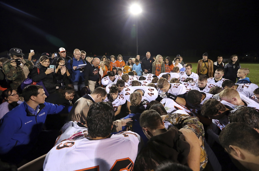 FILE - Bremerton assistant football coach Joe Kennedy, obscured at center in blue, is surrounded by Centralia High School football players as they kneel and pray with him on the field after their game against Bremerton on Oct. 16, 2015, in Bremerton, Wash. After losing his coaching job for refusing to stop kneeling in prayer with players and spectators on the field immediately after football games, Kennedy will take his arguments before the U.S. Supreme Court on Monday, April 25, 2022, saying the Bremerton School District violated his First Amendment rights by refusing to let him continue praying at midfield after games. (Meegan M.
