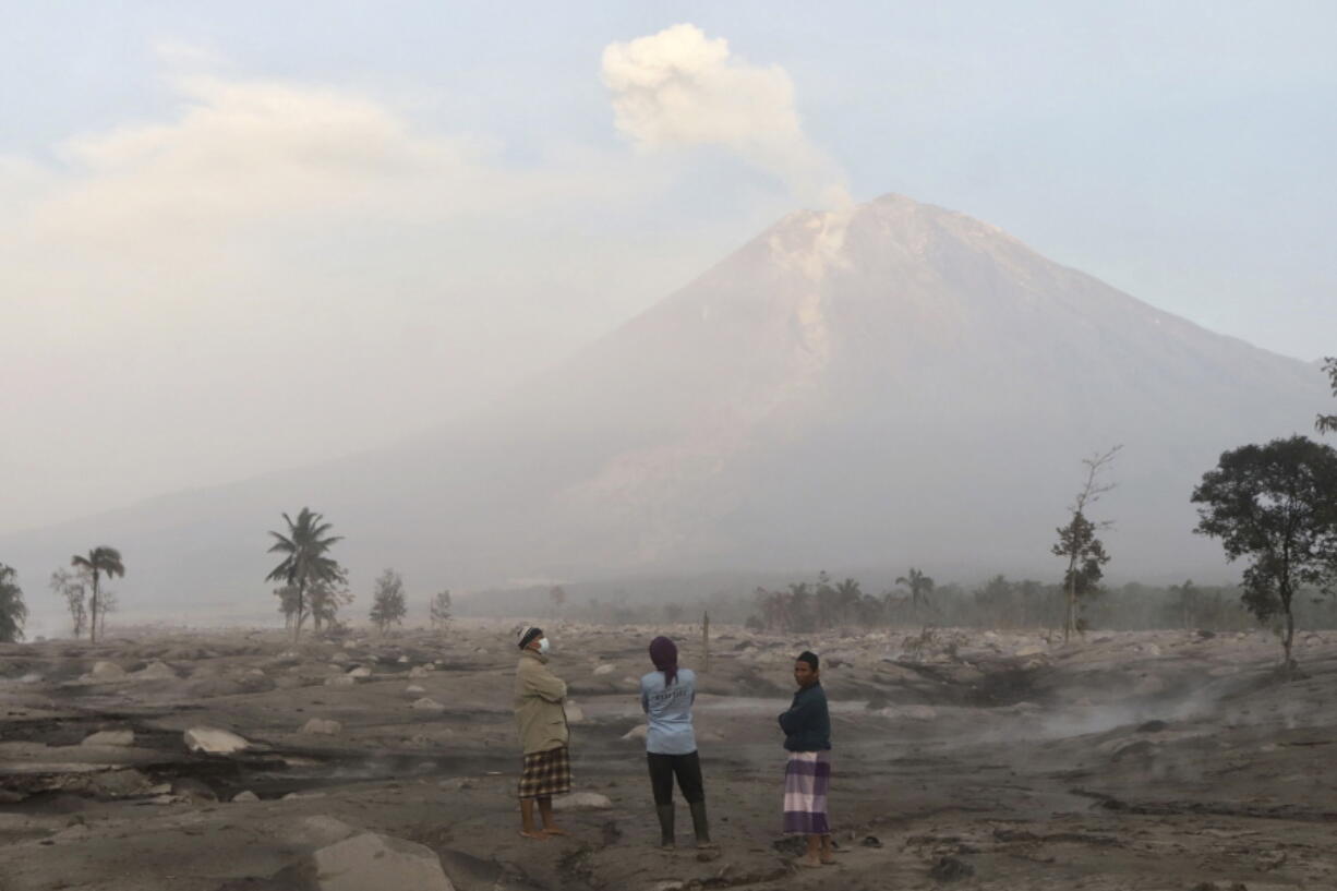 Villagers stand on an area covered in volcanic ash as Mount Semeru looms in the background in Kajar Kuning village in Lumajang, East Java, Indonesia, Monday, Dec. 5, 2022. Indonesia's highest volcano on its most densely populated island released searing gas clouds and rivers of lava Sunday in its latest eruption.