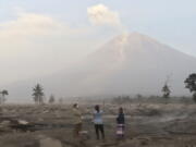 Villagers stand on an area covered in volcanic ash as Mount Semeru looms in the background in Kajar Kuning village in Lumajang, East Java, Indonesia, Monday, Dec. 5, 2022. Indonesia's highest volcano on its most densely populated island released searing gas clouds and rivers of lava Sunday in its latest eruption.