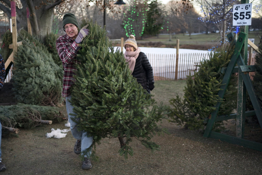 Larry Gurnee carries a $55 Christmas tree he selected with his wife, , Libby Gurnee, background, at a Rotary Club tree sale, Wednesday, Dec. 14, 2022, in South Portland, Maine. Inflation has Americans cutting back on spending in some areas this holiday season but Christmas trees is not one of them, according to the National Christmas Tree Association. (AP Photo/Robert F.
