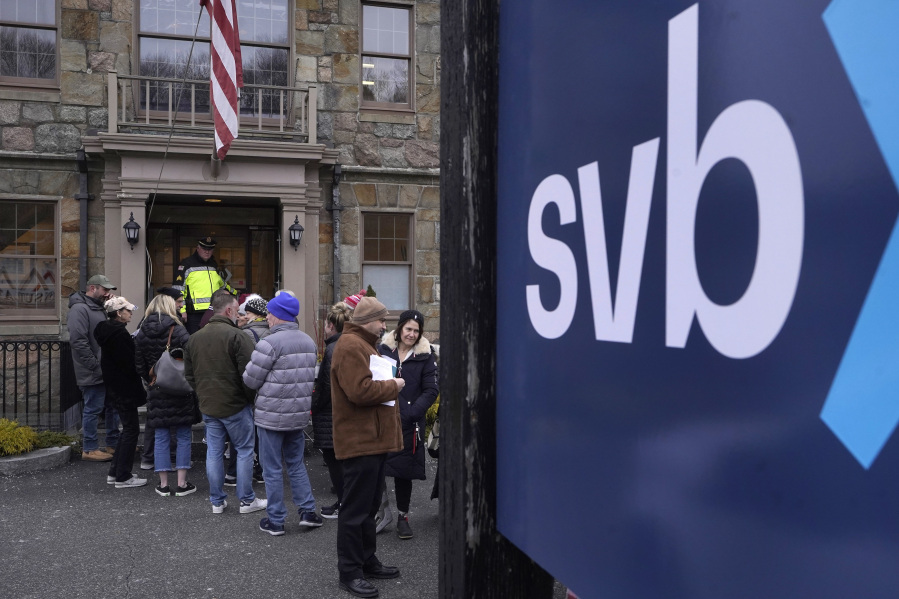 A law enforcement official, behind, stands in an entryway to a Silicon Valley Bank branch location, Monday, March 13, 2023, as customers and bystanders line up outside the bank, in Wellesley, Mass.