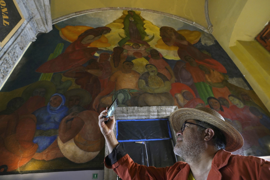 A tourist takes a photo backdropped by the "Alegoria de la Virgen de Guadalupe" mural, in the main entrance of the Antiguo Colegio de San Ildefonso, in Mexico City, Wednesday, April 26, 2023. The mural was created by Mexican artist Fermin Revueltas between 1922 and 1923, when the walls of San Ildefonso became the canvases where the muralist movement came to life.