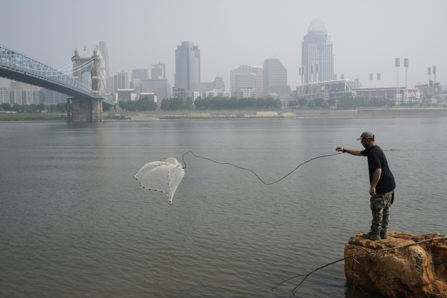 FILE - A man tosses a fishing net into the Ohio River as smoke from wildfires is visible over downtown Cincinnati, June 28, 2023. Forecasters say there won't be large breaks for much of America anytime soon from eye-watering dangerous smoke from fire-struck Canada. (AP Photo/Joshua A.