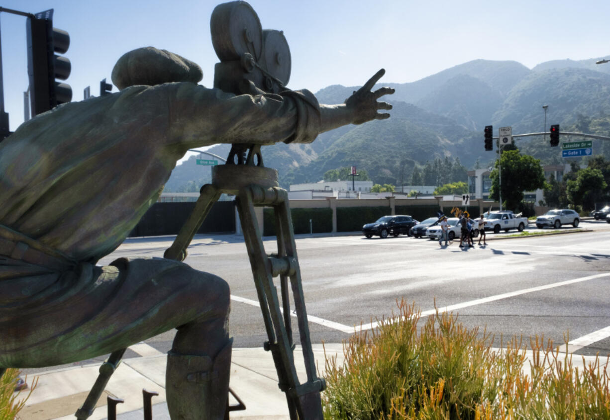 With a statue of a cameraman in the foreground, SAG-AFTRA picketers carrying signs cross a street near the gates of Warner Bros. studios in Burbank, Calif., Tuesday, Sep. 26, 2023.