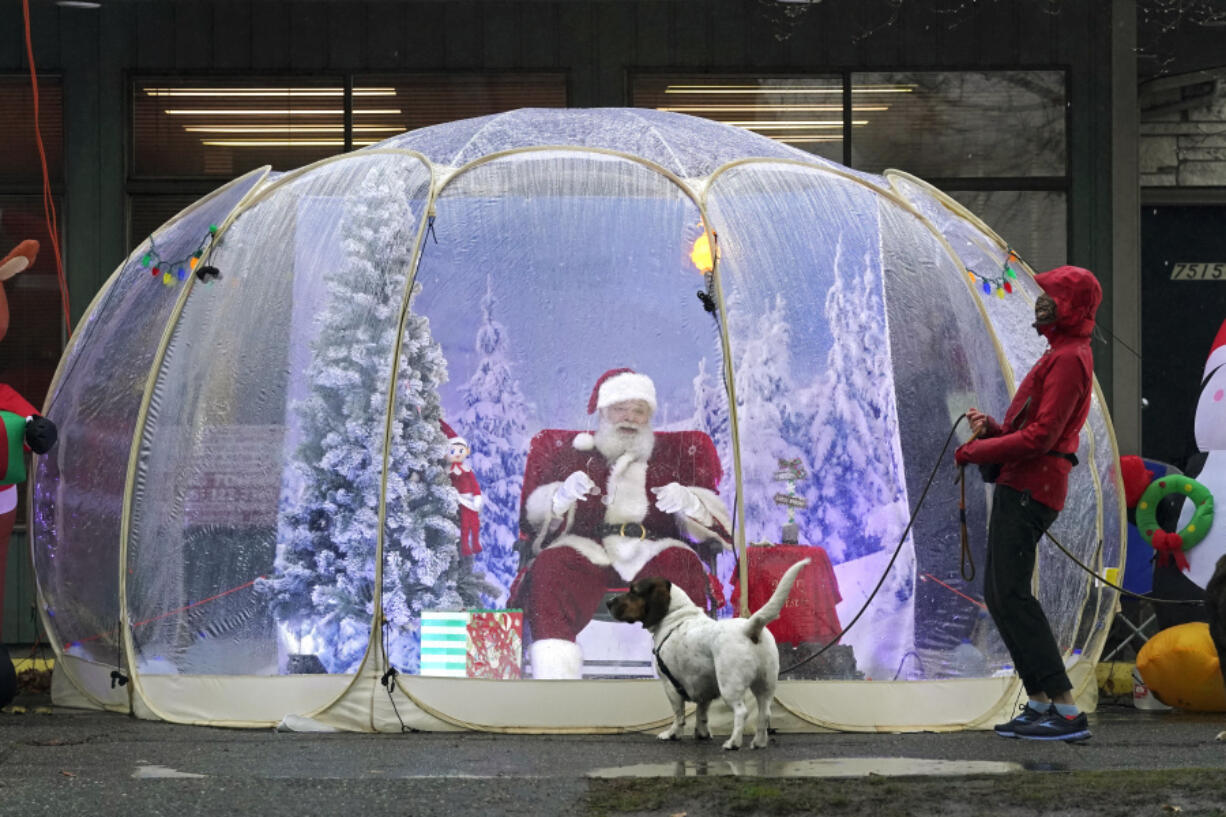 FILE - Santa Claus, portrayed by Dan Kemmis, laughs as he talks to Kristin Laidre as she walks her dog, Scooby, a Bassett Hound mix, as he sits inside a &ldquo;snow globe&rdquo; protective bubble due to the coronavirus pandemic in Seattle&rsquo;s Greenwood neighborhood on Dec. 8, 2020. (AP Photo/Ted S.