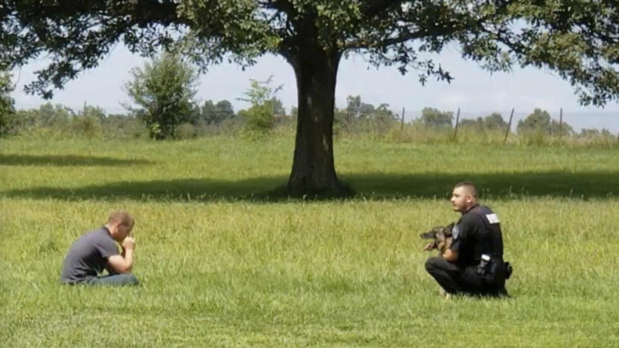 In this image from video provided by a family friend, Taylor Ware, left, sits in a field approached by a police officer and canine at a highway rest stop in Dale, Ind., on Aug. 25, 2019. Taylor's mother called 911 when he wouldn’t get back in their SUV during a manic episode caused by bipolar disorder.